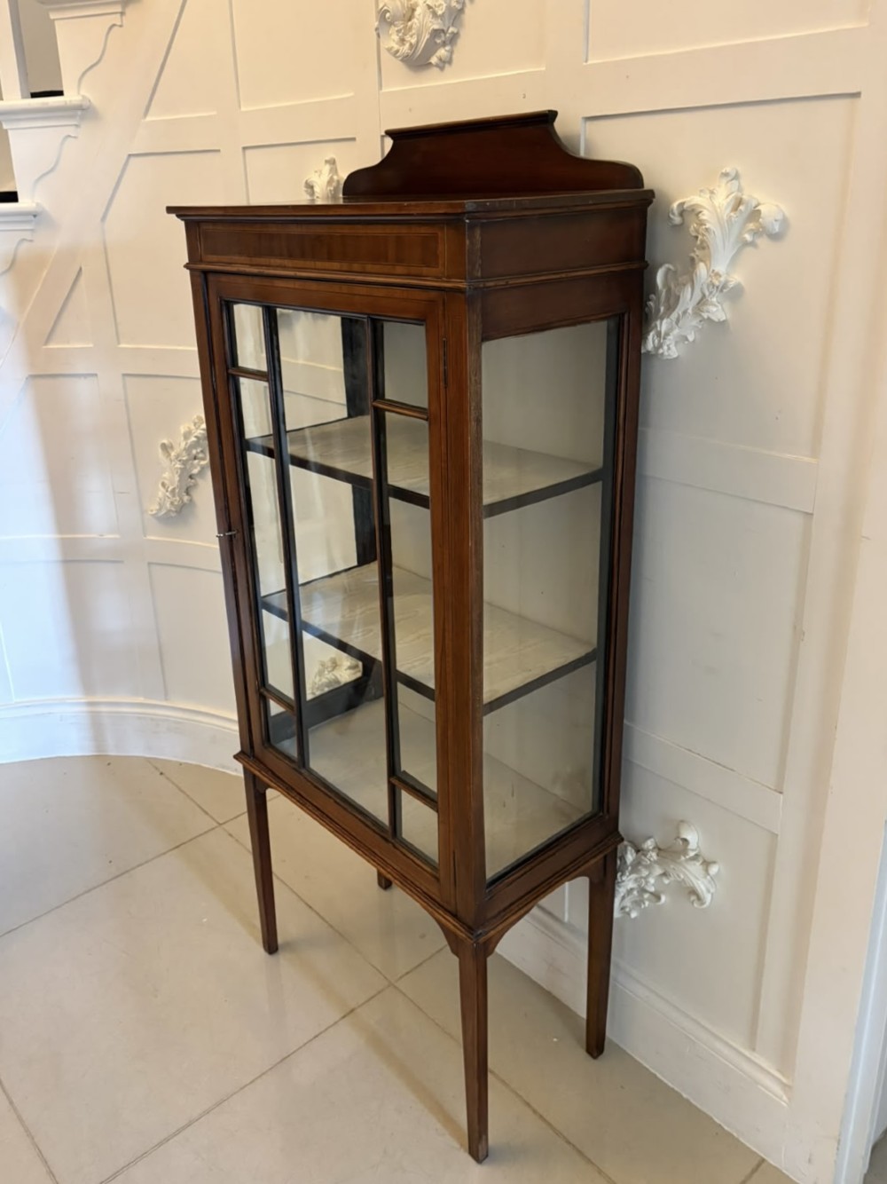 An Edwardian mahogany glazed display cabinet featuring geometric glazing bars, tapered legs, a raised pediment top, and a light-lined interior with three shelves. View VI
