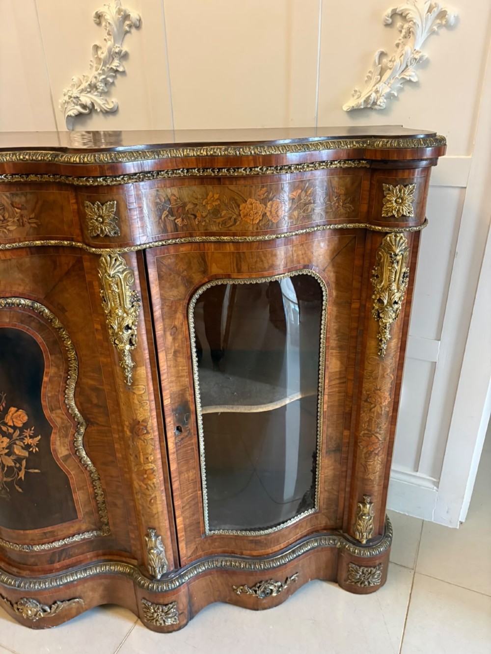 A Victorian serpentine burr walnut credenza featuring detailed floral marquetry inlays, gilt-bronze mounts, a central panelled door, and two curved glazed side doors. View XII