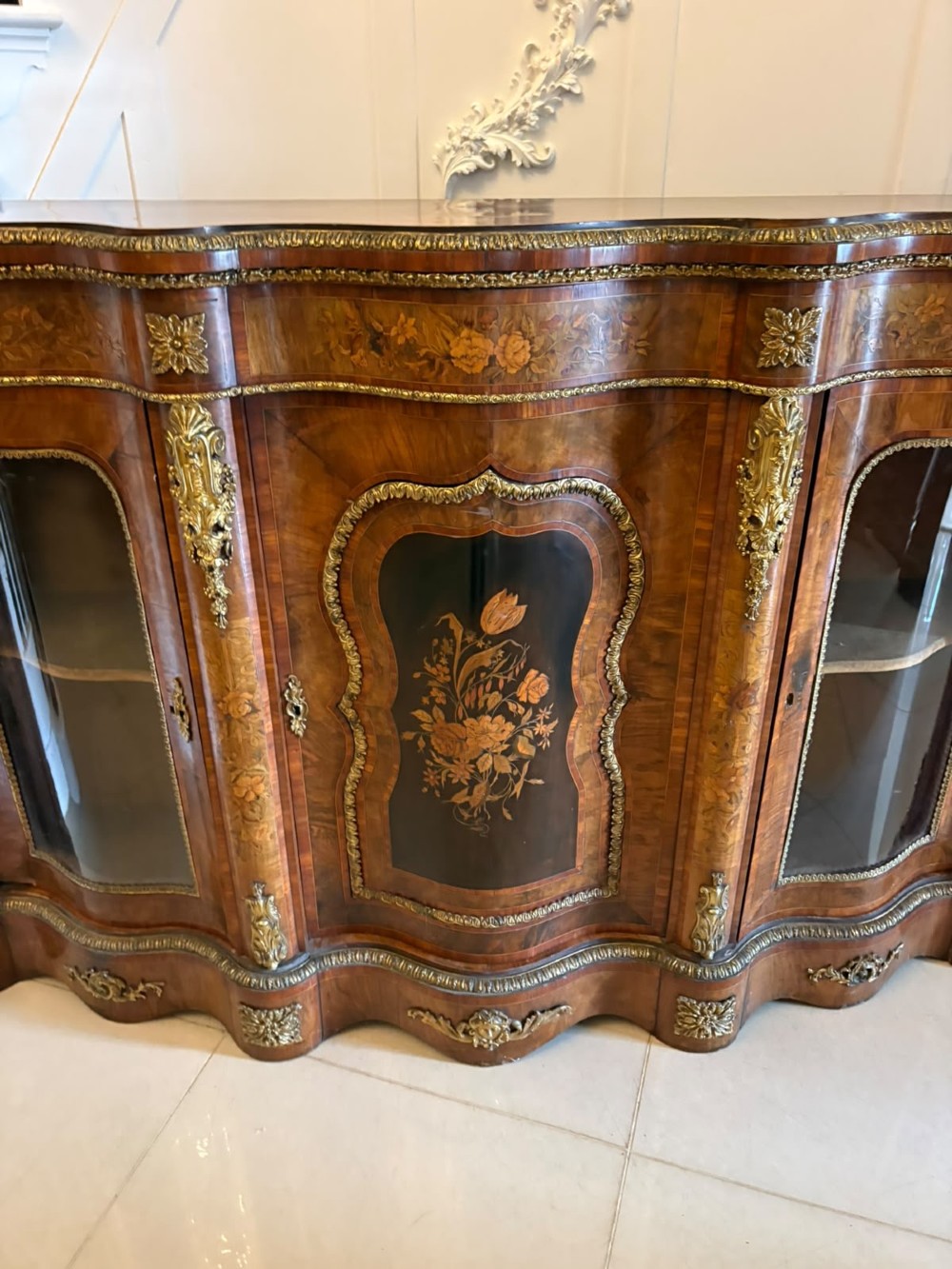 A Victorian serpentine burr walnut credenza featuring detailed floral marquetry inlays, gilt-bronze mounts, a central panelled door, and two curved glazed side doors. View XIV