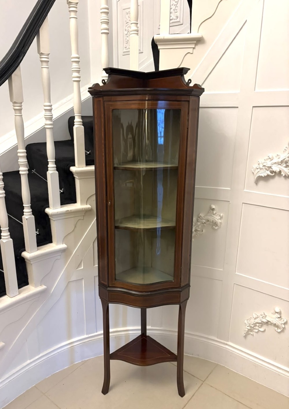 An Edwardian mahogany corner display cabinet with a serpentine glazed door, line inlay decoration, shaped upper pediment, and slender tapered legs with a lower undertier.