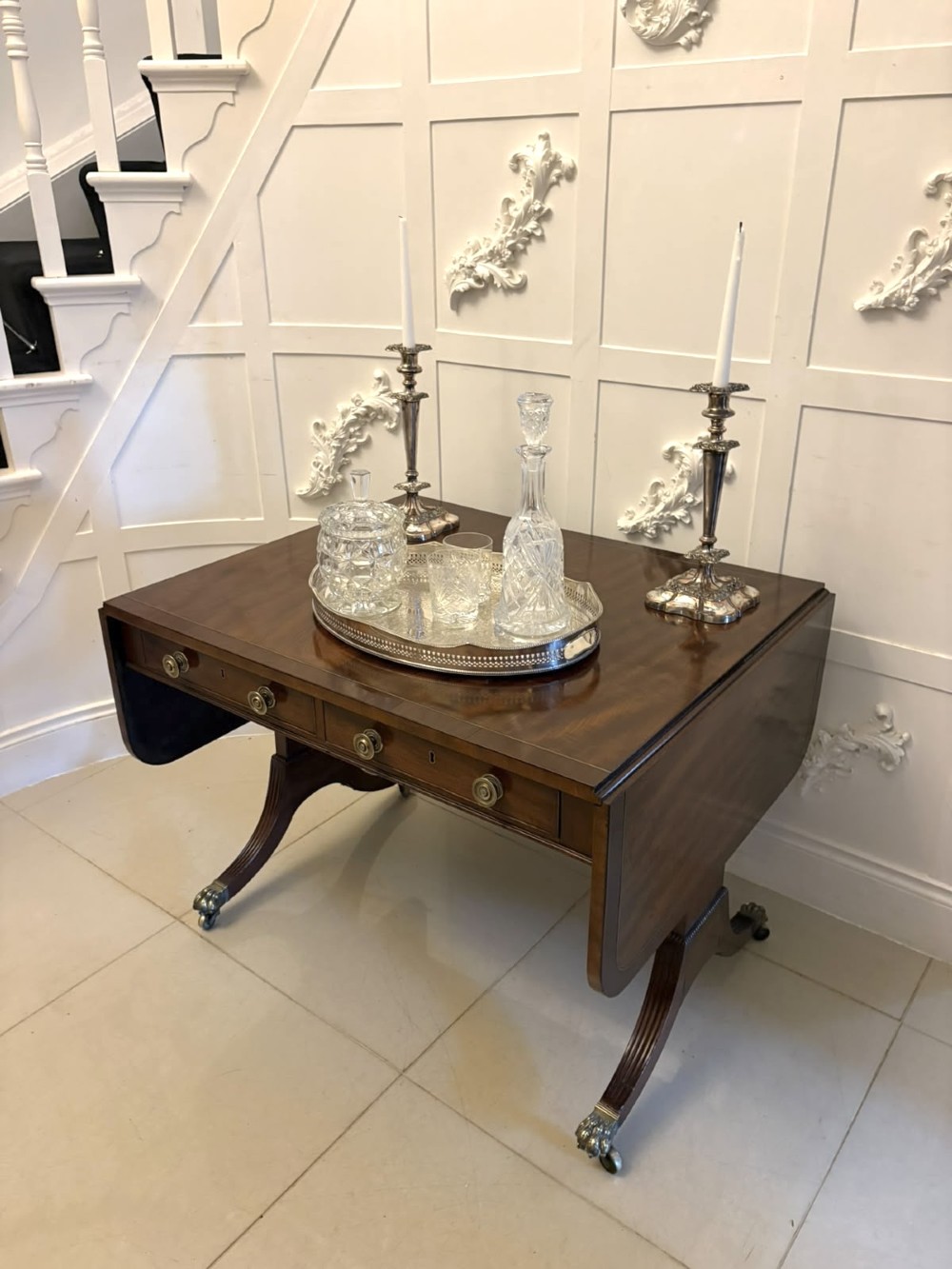 A Regency mahogany sofa table featuring two drop leaves, two front drawers with round brass knobs, reeded splayed legs, and brass paw castors. View III