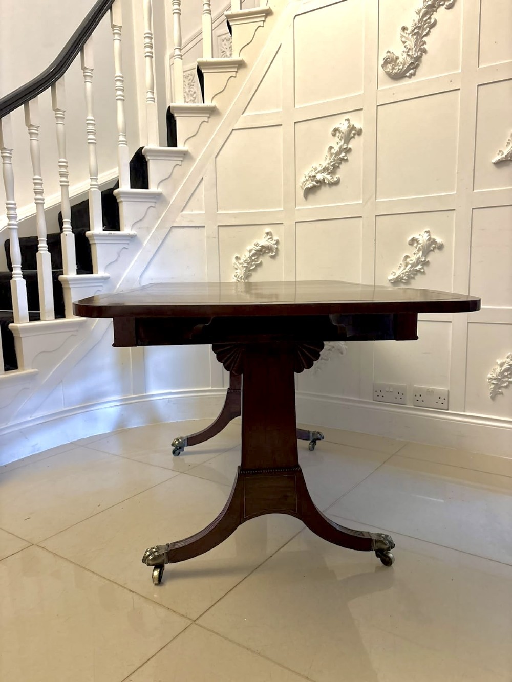 A Regency mahogany sofa table featuring two drop leaves, two front drawers with round brass knobs, reeded splayed legs, and brass paw castors. View VI