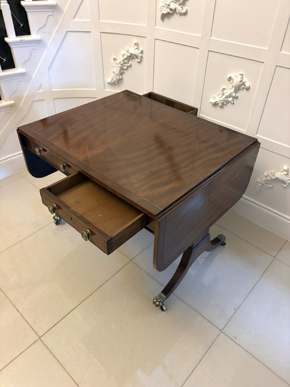 A Regency mahogany sofa table featuring two drop leaves, two front drawers with round brass knobs, reeded splayed legs, and brass paw castors. View VIII