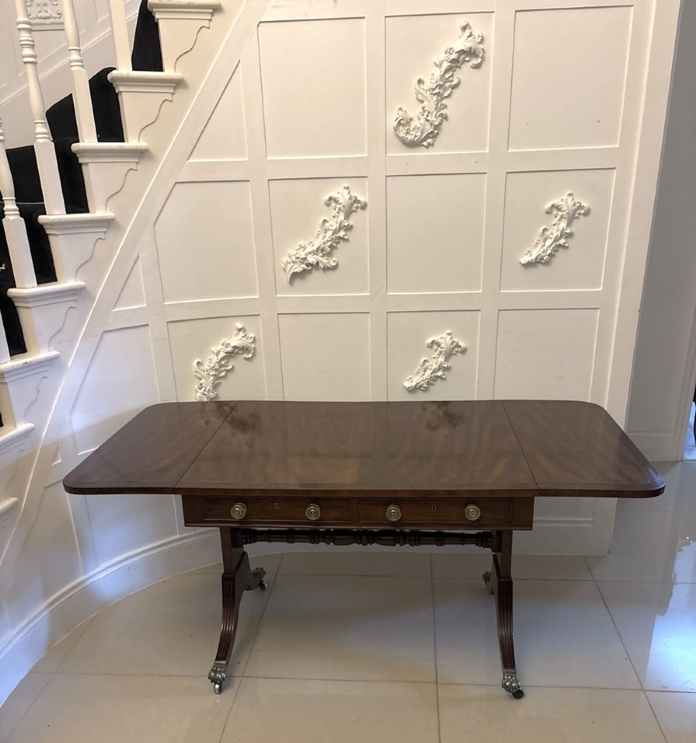 A Regency mahogany sofa table featuring two drop leaves, two front drawers with round brass knobs, reeded splayed legs, and brass paw castors. View IX