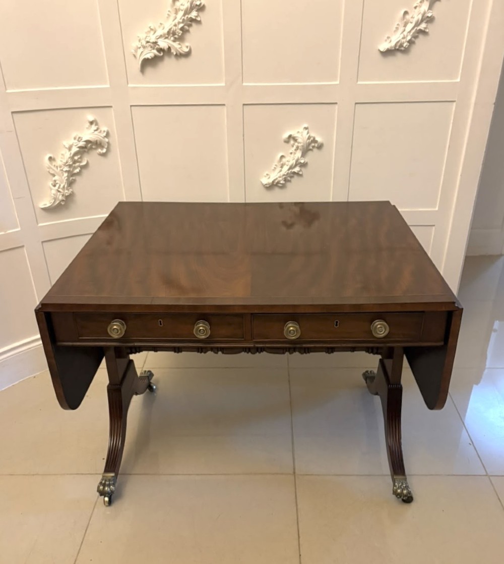 A Regency mahogany sofa table featuring two drop leaves, two front drawers with round brass knobs, reeded splayed legs, and brass paw castors.