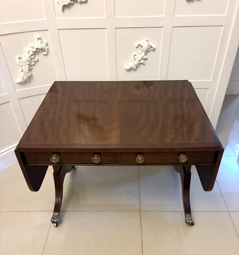 A Regency mahogany sofa table featuring two drop leaves, two front drawers with round brass knobs, reeded splayed legs, and brass paw castors. View XV