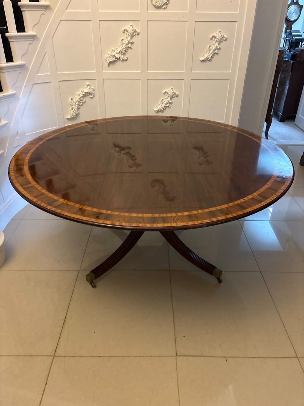 A large circular mahogany dining table featuring rosewood and satinwood crossbanding set upon a central pedestal base with reeded legs ending in brass castors.