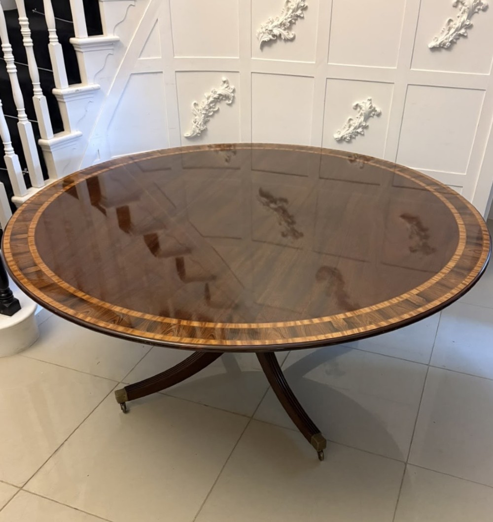 A large circular mahogany dining table featuring rosewood and satinwood crossbanding set upon a central pedestal base with reeded legs ending in brass castors. View XII