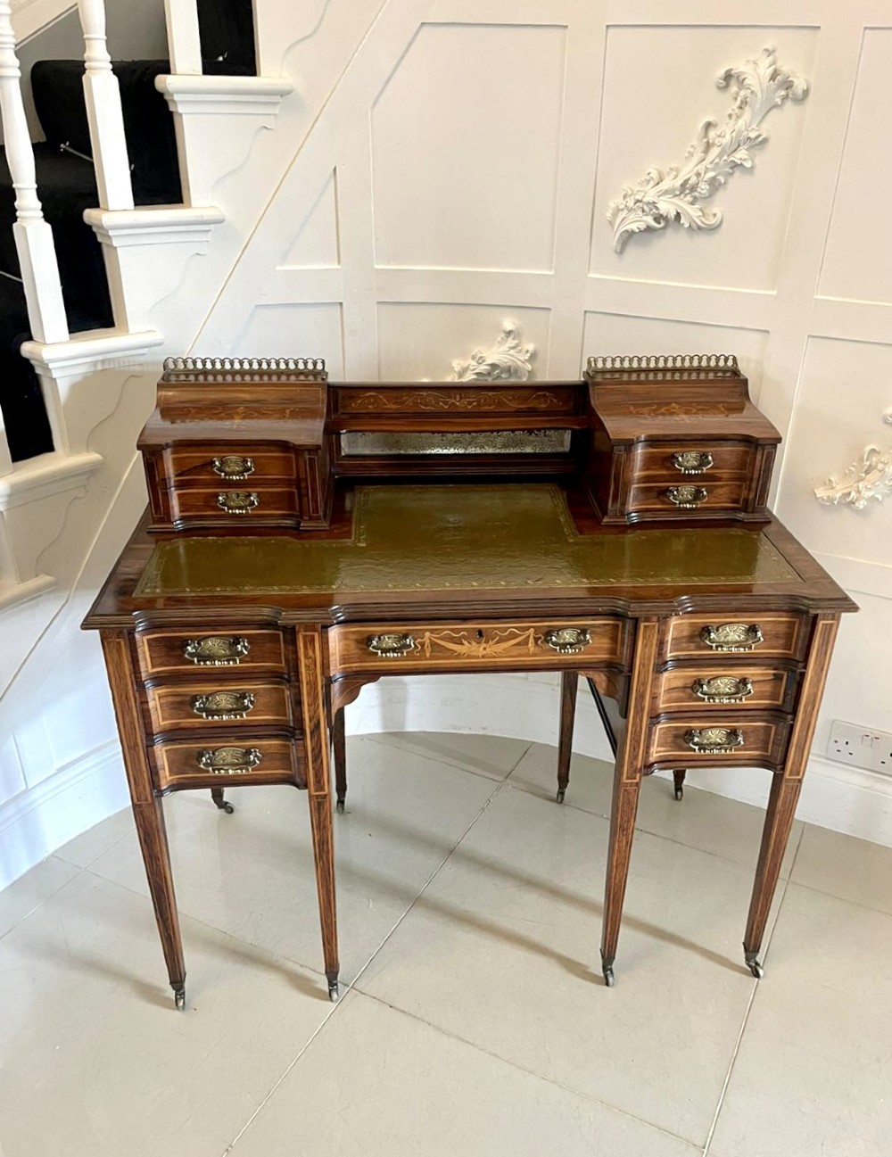 A late Victorian inlaid writing desk with a dark rosewood veneer, green leather top insert, brass gallery rail, and multiple drawers on tapered legs.