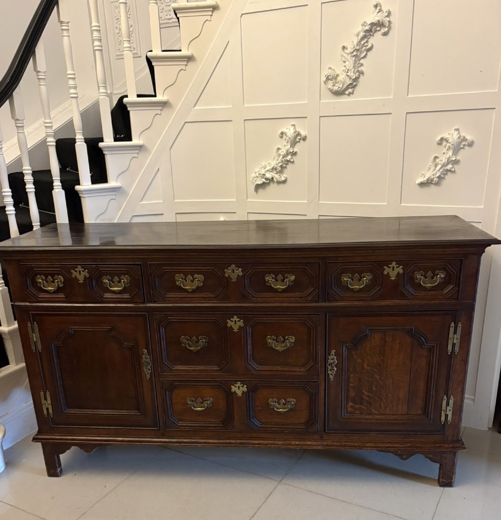 An antique dark oak Georgian dresser base featuring geometric panel drawers, brass handles, side cupboard doors, and a smooth wooden surface.
