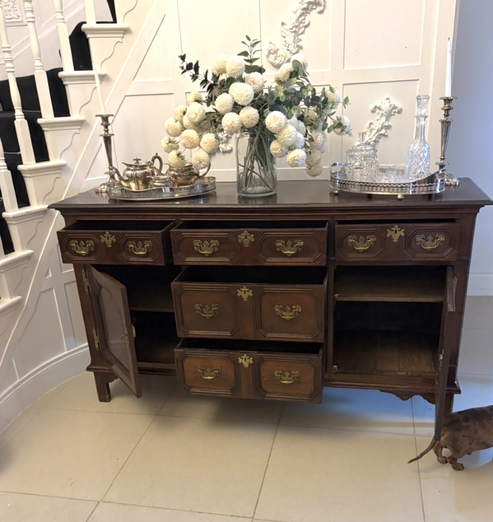 An antique dark oak Georgian dresser base featuring geometric panel drawers, brass handles, side cupboard doors, and a smooth wooden surface. View XII