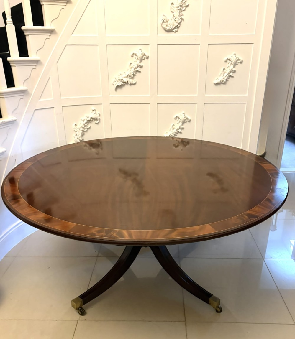 A circular mahogany dining table with a crossbanded edge tilted vertically on a central pedestal base with splayed legs and brass castors. View VI