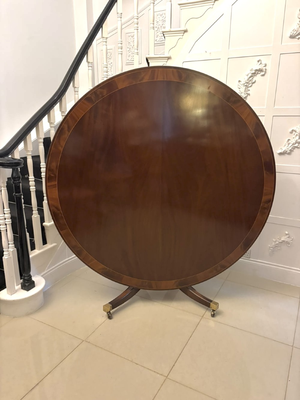 A circular mahogany dining table with a crossbanded edge tilted vertically on a central pedestal base with splayed legs and brass castors.