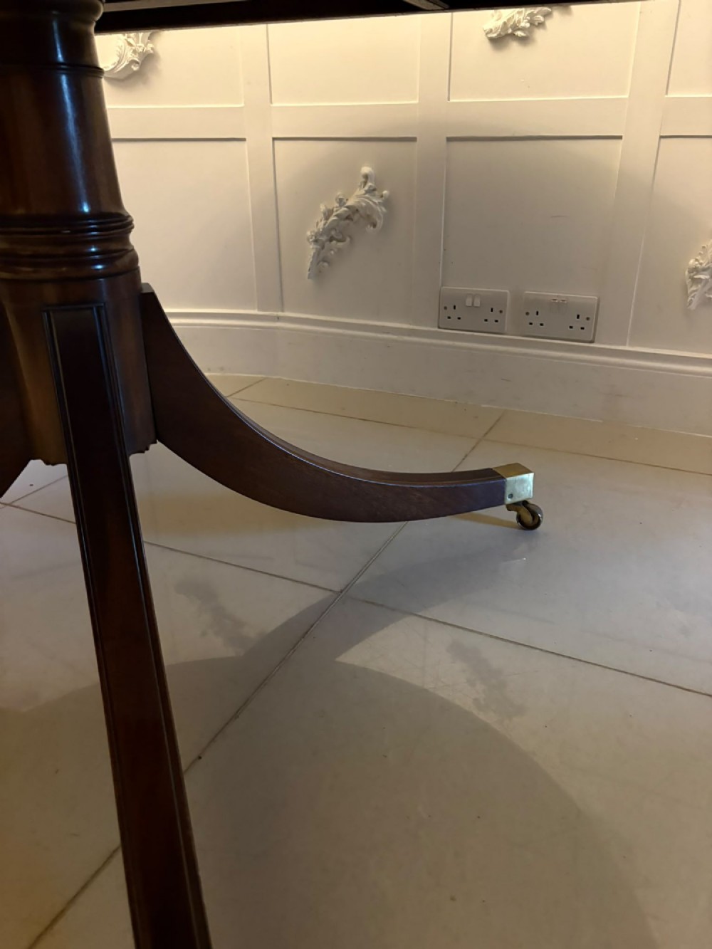A circular mahogany dining table with a crossbanded edge tilted vertically on a central pedestal base with splayed legs and brass castors. View IX
