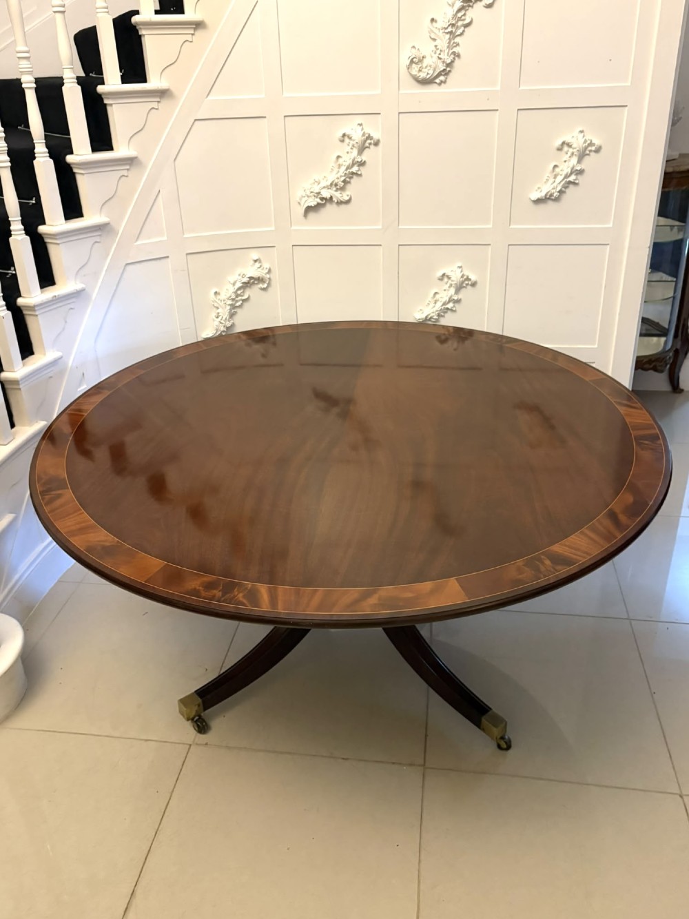A circular mahogany dining table with a crossbanded edge tilted vertically on a central pedestal base with splayed legs and brass castors. View X