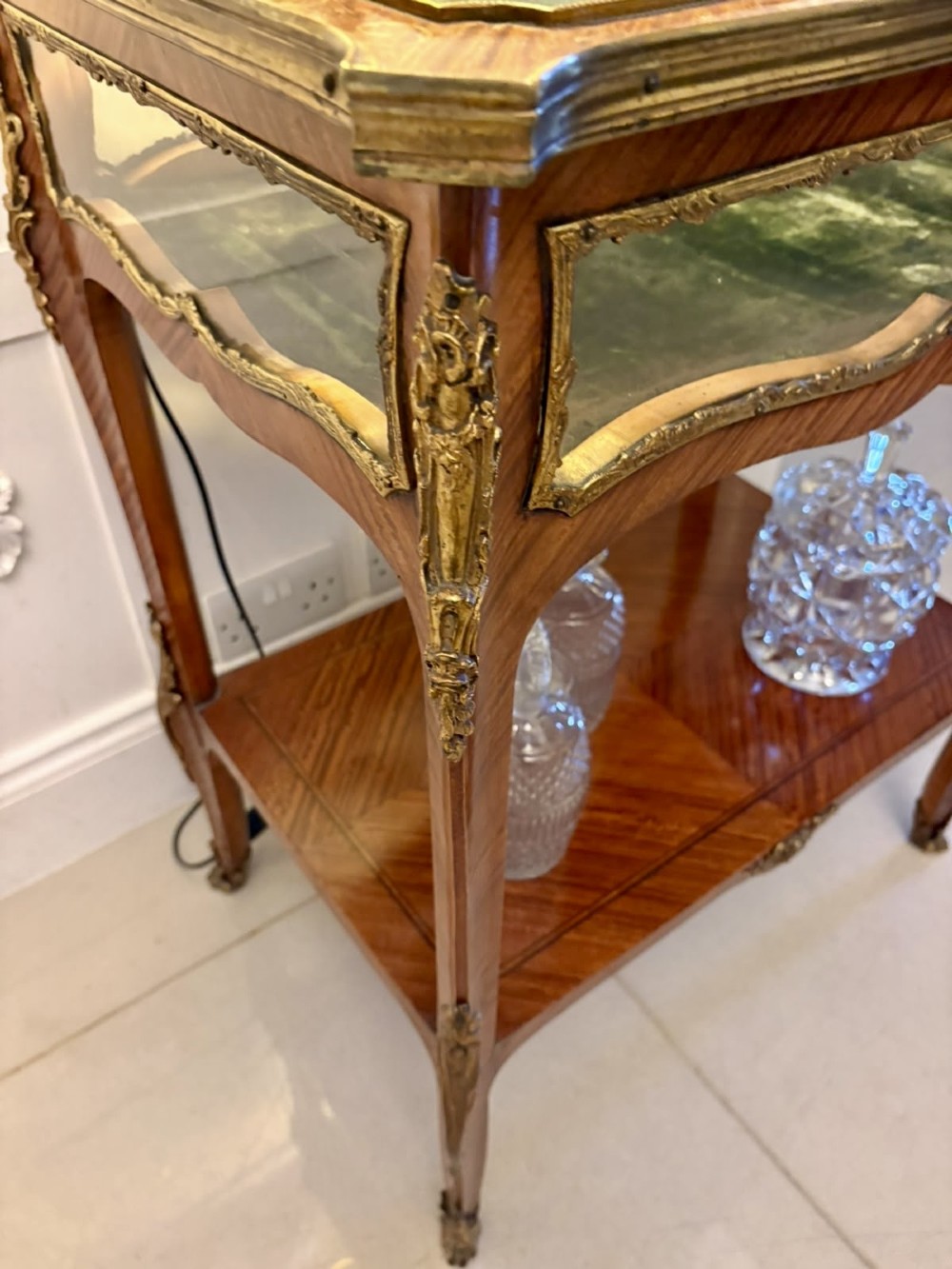 An antique French Napoleon III satinwood bijouterie table featuring a glazed display top, curved cabriole legs with decorative brass mounts, and an undertier shelf. View X