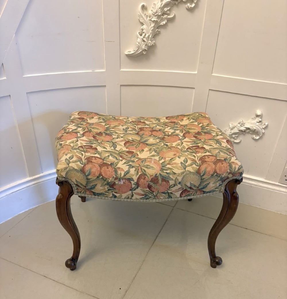 A low rectangular Victorian footstool featuring carved walnut cabriole legs and a padded seat upholstered in a botanical tapestry fabric depicting fruit and foliage. View X