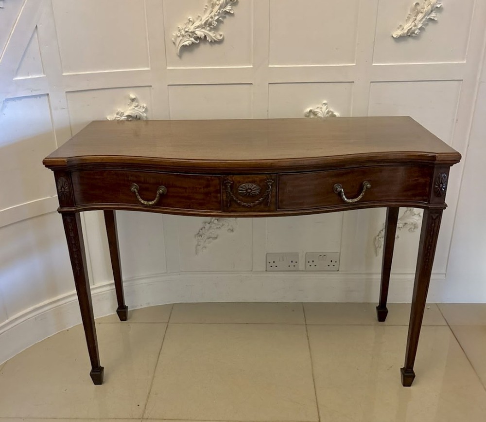 An Edwardian mahogany console table featuring a serpentine front, carved central motif, brass handles, and square tapered legs.