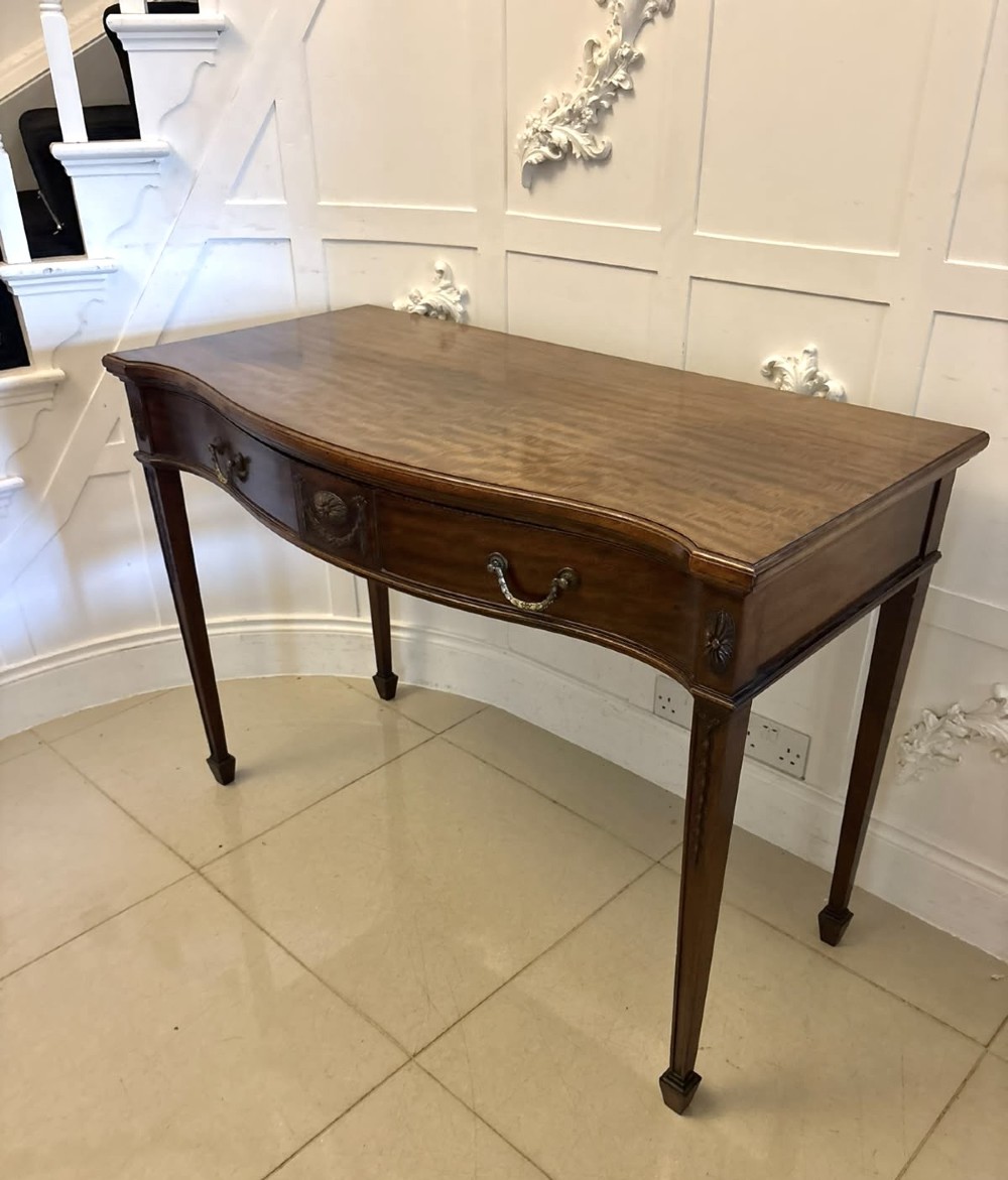 An Edwardian mahogany console table featuring a serpentine front, carved central motif, brass handles, and square tapered legs. View X