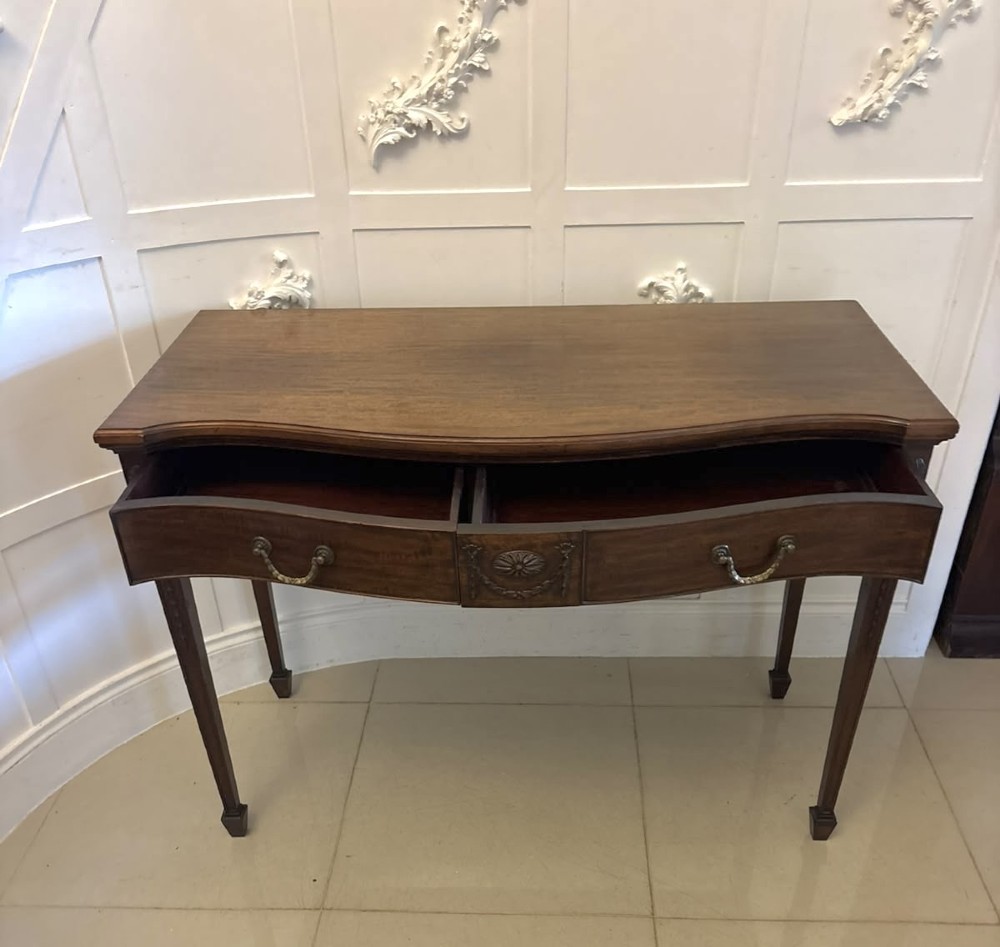 An Edwardian mahogany console table featuring a serpentine front, carved central motif, brass handles, and square tapered legs. View XI