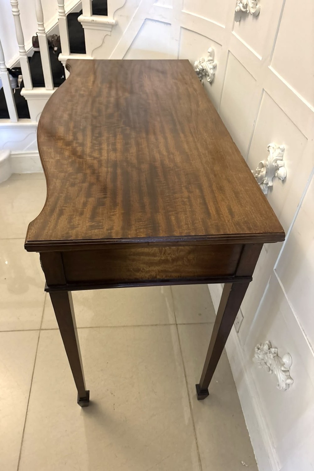An Edwardian mahogany console table featuring a serpentine front, carved central motif, brass handles, and square tapered legs. View XII