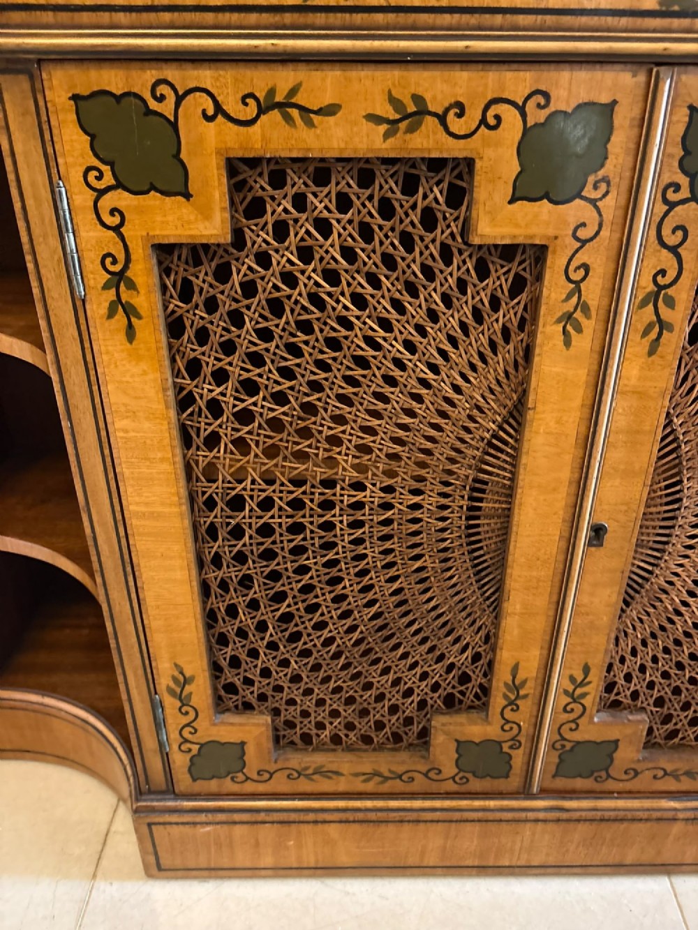 A Sheraton Revival satinwood side cabinet featuring painted leaf borders, central doors with fan-shaped woven cane panels, and open curved side shelves. View XI