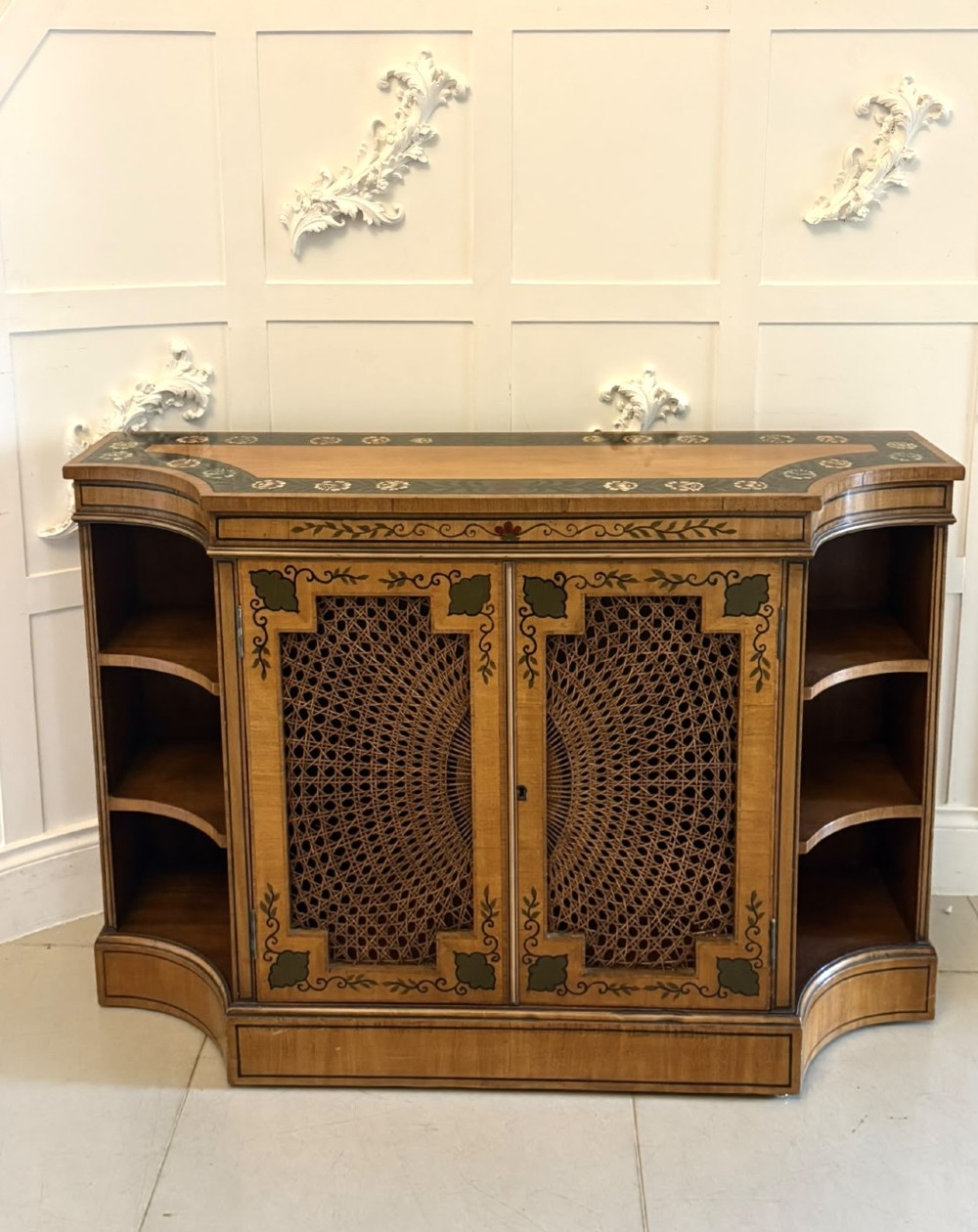 A Sheraton Revival satinwood side cabinet featuring painted leaf borders, central doors with fan-shaped woven cane panels, and open curved side shelves.