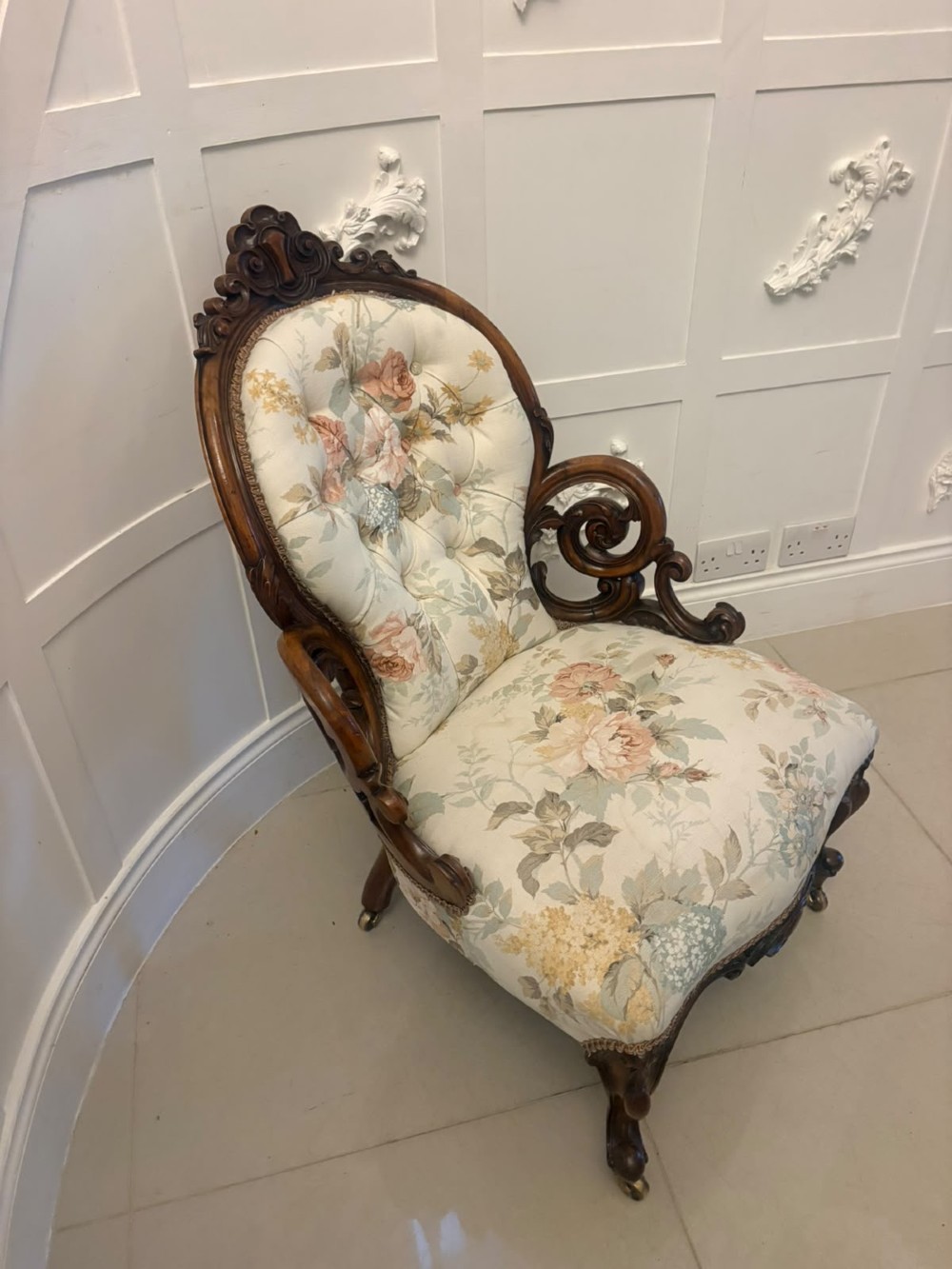 A Victorian walnut armchair featuring floral button-tufted upholstery, carved open scroll arms, and front legs set on brass castors.