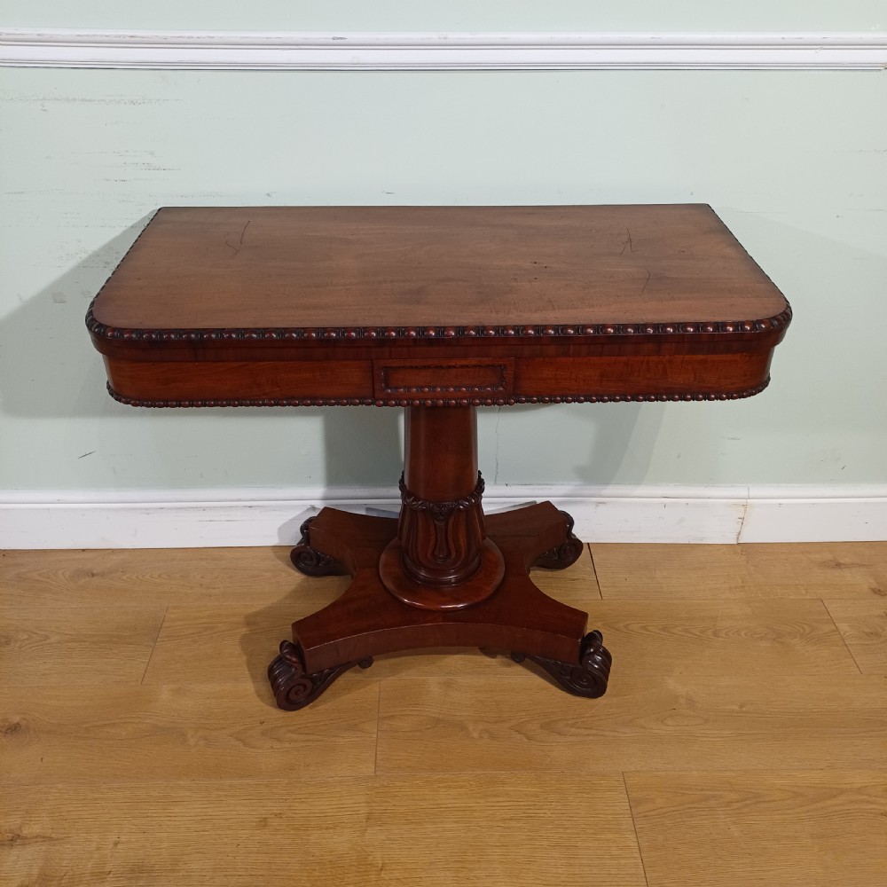 A nineteenth-century mahogany fold-over card table featuring a rectangular top with rounded corners, bead moulding along the frieze, a turned pedestal column, and a quadripartite base with carved scroll feet.