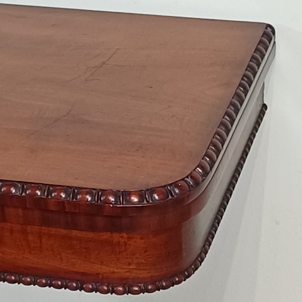 A nineteenth-century mahogany fold-over card table featuring a rectangular top with rounded corners, bead moulding along the frieze, a turned pedestal column, and a quadripartite base with carved scroll feet. View X