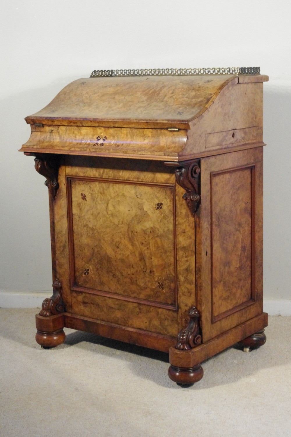 A Victorian burr walnut Davenport desk featuring a sloped hinged writing lid with brass gallery rail, carved scroll supports, inlaid motifs, and bun feet.