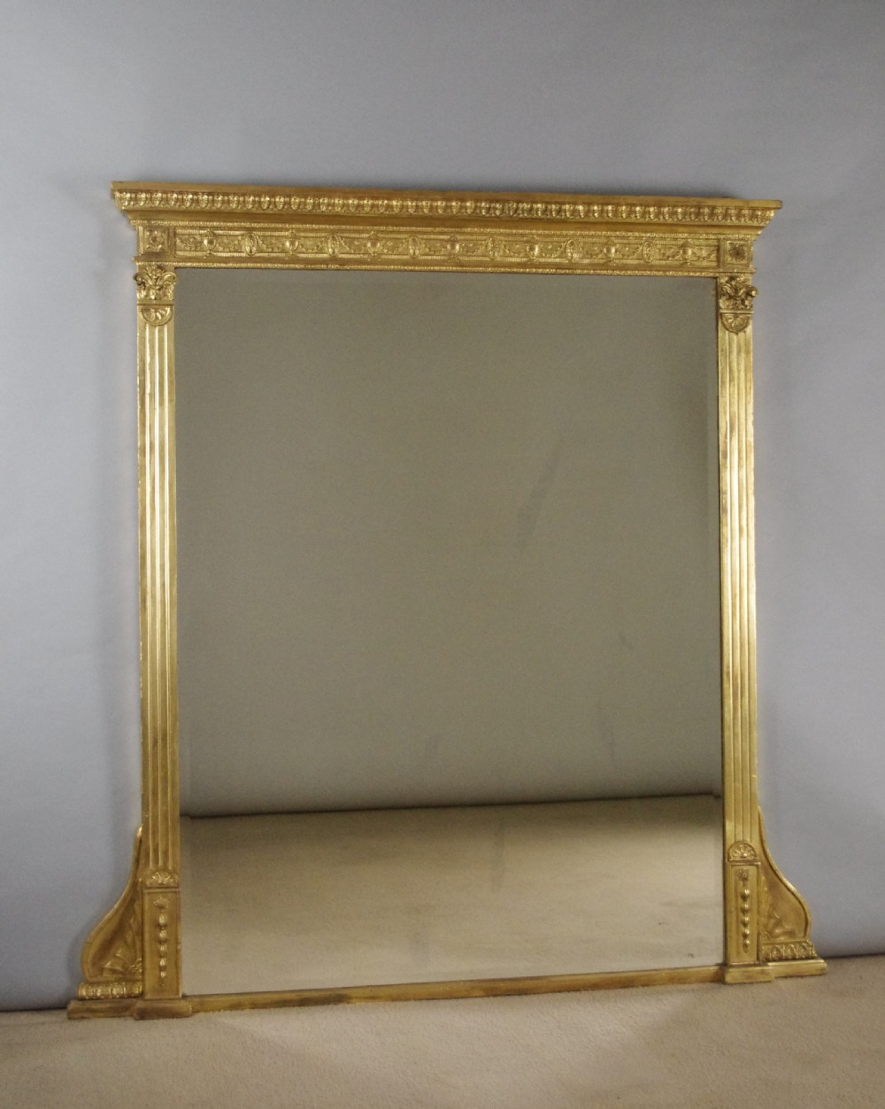 A large rectangular giltwood overmantel mirror in the Neoclassical style featuring fluted pilasters, classical frieze moulding, and decorative base scrolls.