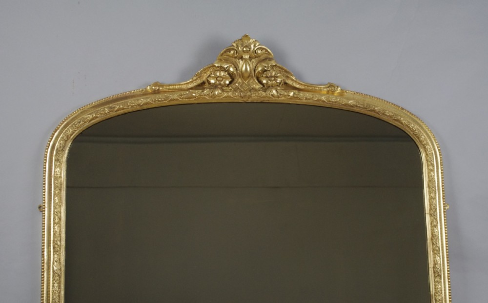 A large Victorian arched gilt overmantle mirror featuring an ornate central floral crest and beaded detail along the frame. View II