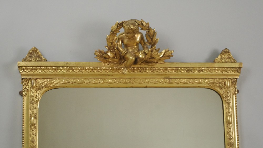 A 19th-century Victorian giltwood overmantel mirror featuring a moulded arch frame decorated with bead moulding and topped with a sculpted putto cresting within a laurel wreath. View II