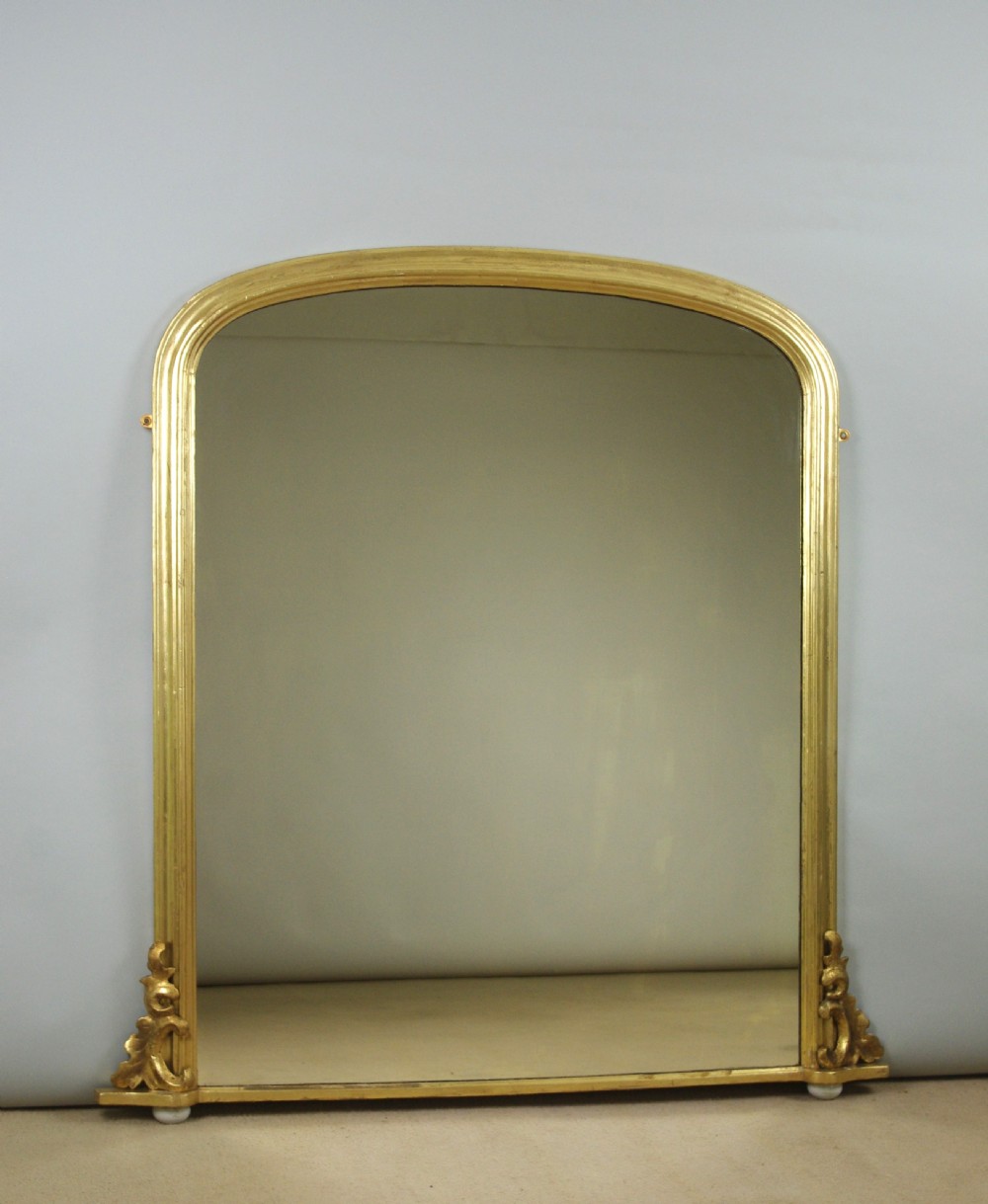 A Victorian arched overmantel mirror featuring a moulded gold-leaf frame with carved scroll flourishes at the lower corners.