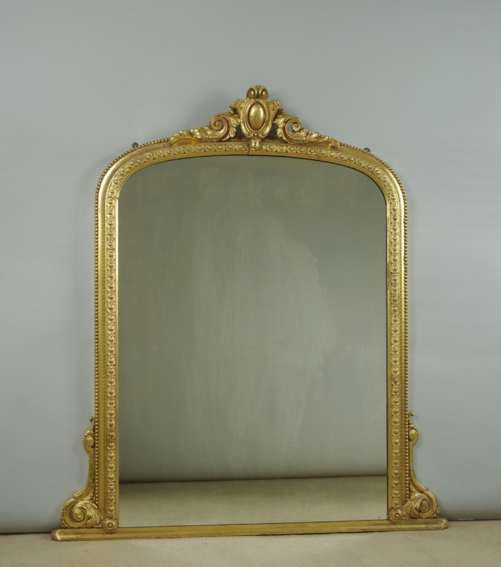 A Victorian giltwood overmantel mirror featuring an arched top decorated with a central crest motif, beaded trim moulding, and side scrolls.
