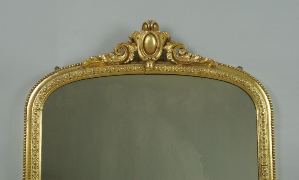 A Victorian giltwood overmantel mirror featuring an arched top decorated with a central crest motif, beaded trim moulding, and side scrolls. View II