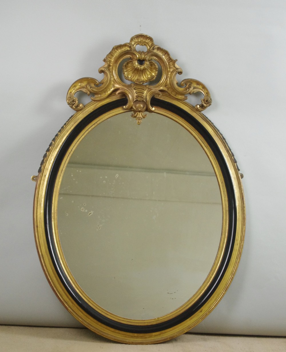 A nineteenth-century French oval wall mirror featuring an ebonised and parcel-gilt frame with a carved shell pediment crest.