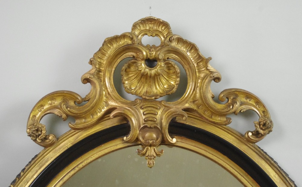 A nineteenth-century French oval wall mirror featuring an ebonised and parcel-gilt frame with a carved shell pediment crest. View II