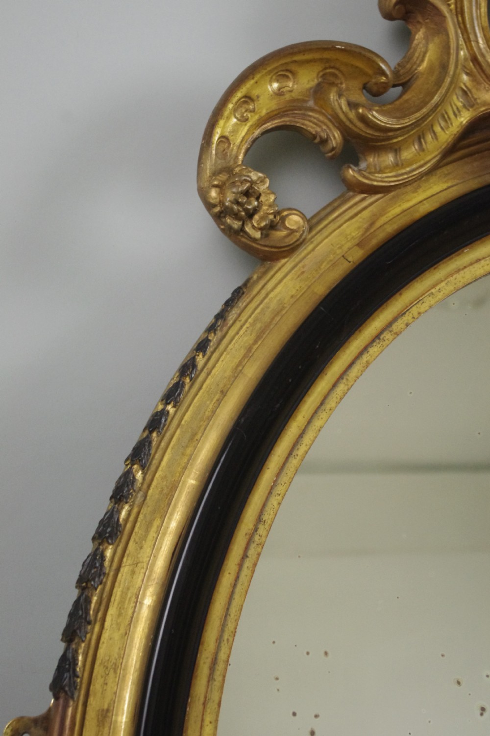 A nineteenth-century French oval wall mirror featuring an ebonised and parcel-gilt frame with a carved shell pediment crest. View IV
