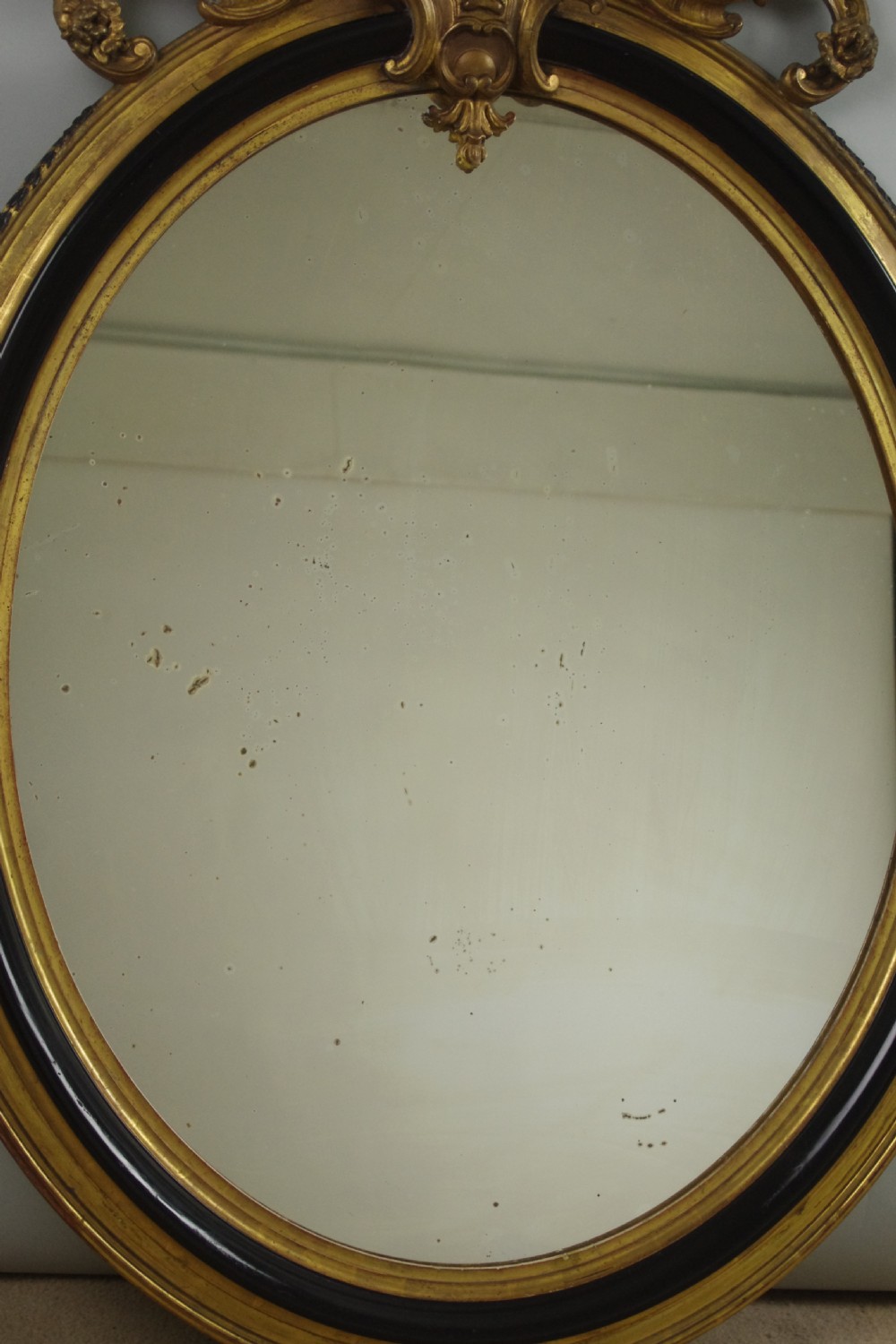 A nineteenth-century French oval wall mirror featuring an ebonised and parcel-gilt frame with a carved shell pediment crest. View IX