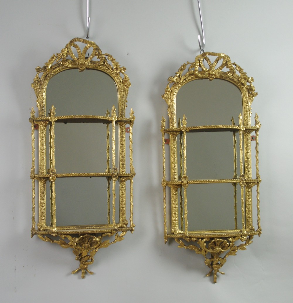 A matching pair of Victorian giltwood wall shelves featuring mirrored backs, ornate bow cresting, and open tiers for displaying small objects.