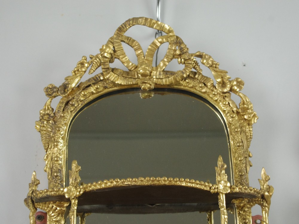 A matching pair of Victorian giltwood wall shelves featuring mirrored backs, ornate bow cresting, and open tiers for displaying small objects. View V