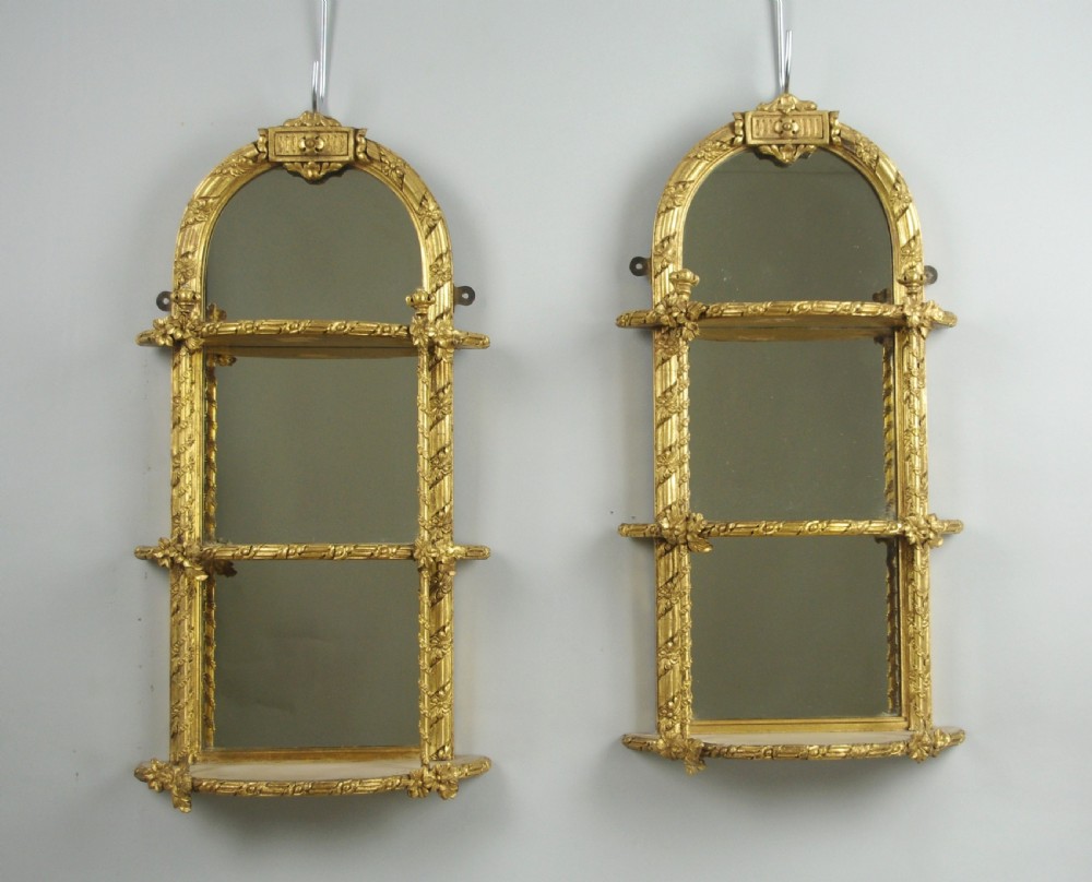 A pair of matching Victorian arched gilt wall mirrors featuring integrated display shelves and ornate moulded frames against a plain background.