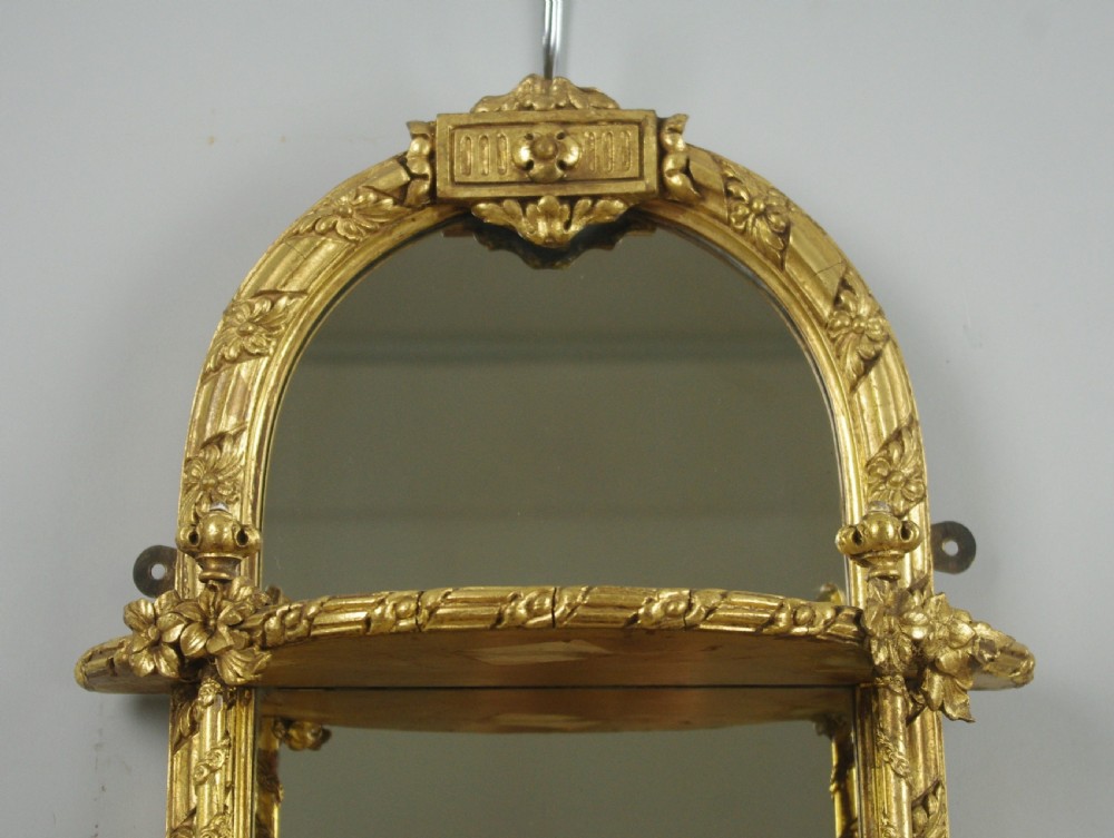 A pair of matching Victorian arched gilt wall mirrors featuring integrated display shelves and ornate moulded frames against a plain background. View IV