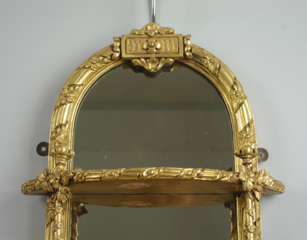 A pair of matching Victorian arched gilt wall mirrors featuring integrated display shelves and ornate moulded frames against a plain background. View V