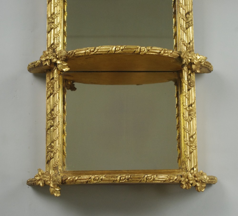 A pair of matching Victorian arched gilt wall mirrors featuring integrated display shelves and ornate moulded frames against a plain background. View VI