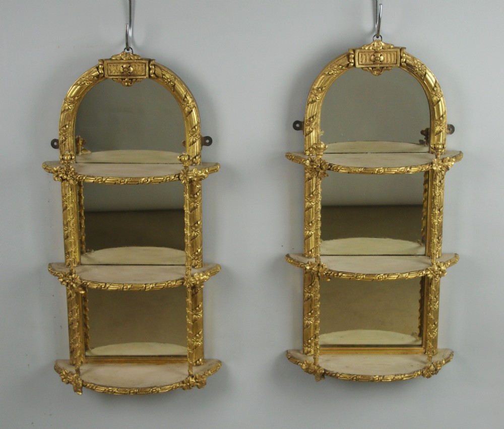 A pair of matching Victorian arched gilt wall mirrors featuring integrated display shelves and ornate moulded frames against a plain background. View X