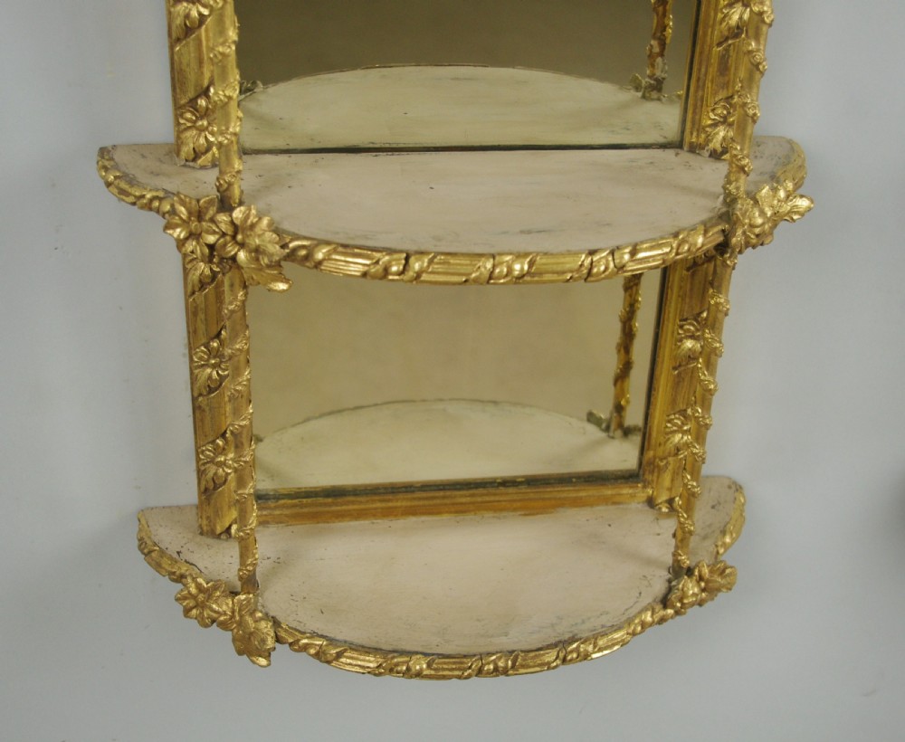 A pair of matching Victorian arched gilt wall mirrors featuring integrated display shelves and ornate moulded frames against a plain background. View XI