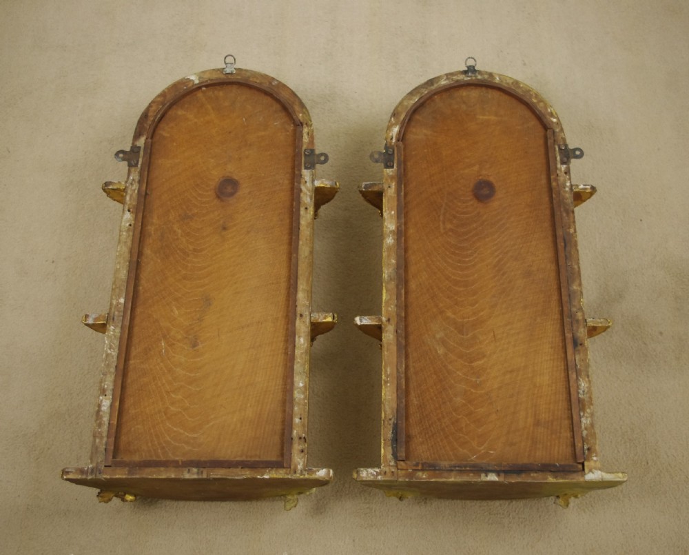 A pair of matching Victorian arched gilt wall mirrors featuring integrated display shelves and ornate moulded frames against a plain background. View XIII